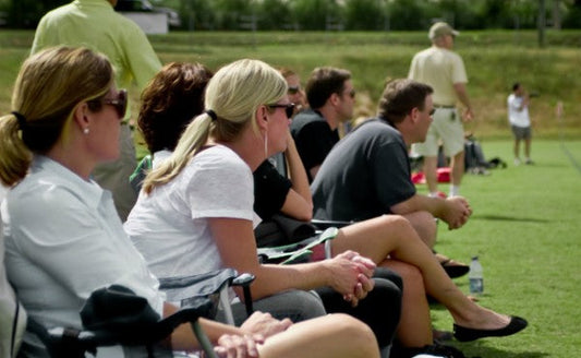 Parents sitting on the sidelines watching a youth lacrosse game on a sunny day in Colorado.